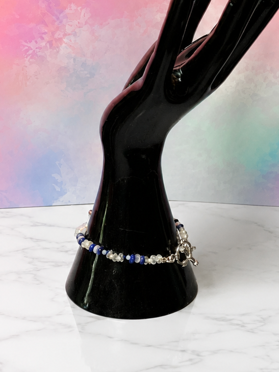 A black hand-shaped jewelry display stand holding a beaded bracelet, set on a white marble surface against a pastel pink, purple, and blue watercolor background.