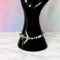 A beaded bracelet with a silver-toned cross charm displayed on a glossy black hand-shaped jewelry stand, set against a pink and blue gradient background.