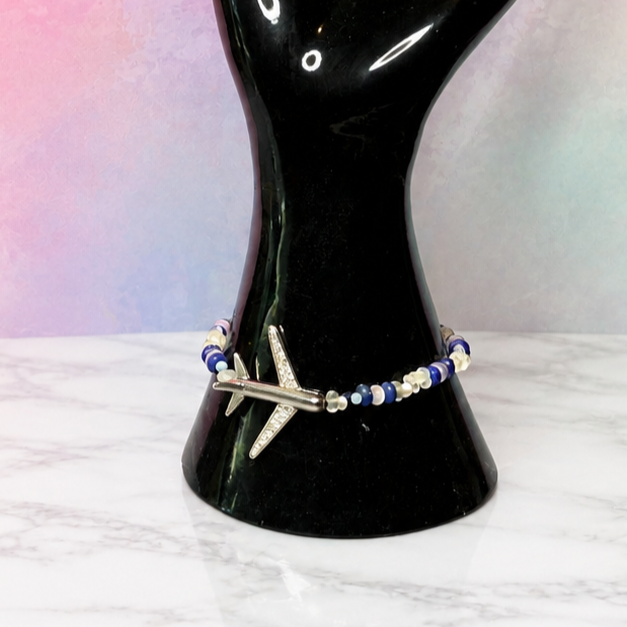 A beaded bracelet with a silver-toned cross charm displayed on a glossy black hand-shaped jewelry stand, set against a pink and blue gradient background.