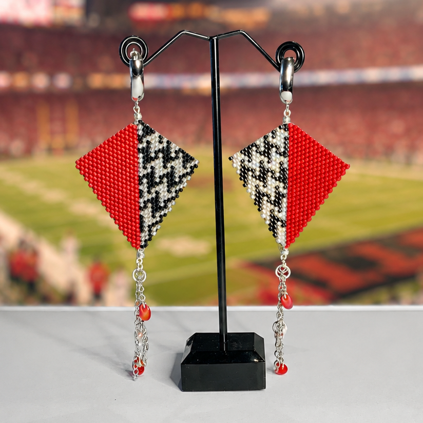 A pair of beaded dangle earrings displayed on a black stand against a blurred stadium background. Each earring has a red and black-and-white houndstooth patterned diamond with dangling red beads and silver-toned chains.