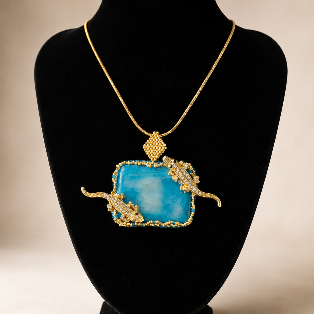 A gold pendant necklace displayed on a black velvet jewelry bust. The pendant features a large, textured light blue rectangular stone with ornate gold side details and a small gold diamond-shaped pendant above it.