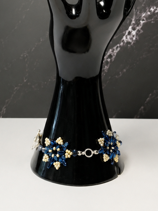 A black glossy hand-shaped jewelry display stand holding a bracelet with blue, white, and gold floral details, set against a dark marble-patterned background and white surface.