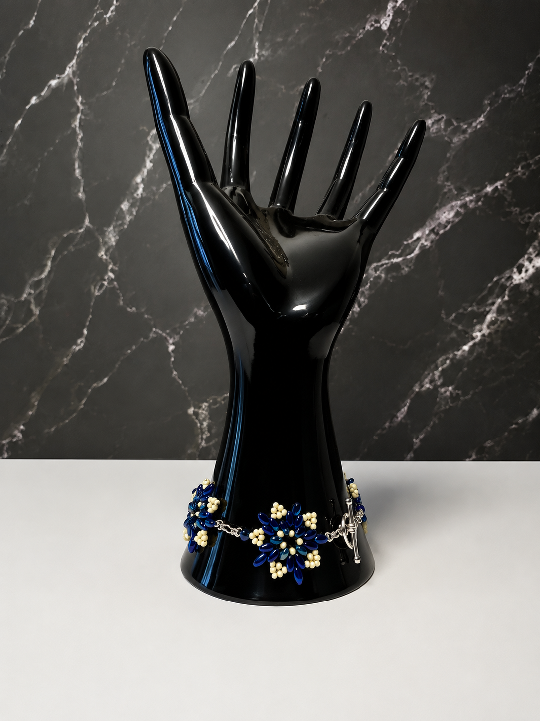 A black hand-shaped jewelry display stand holding a blue and gold floral bracelet, set against a dark marble-patterned background.