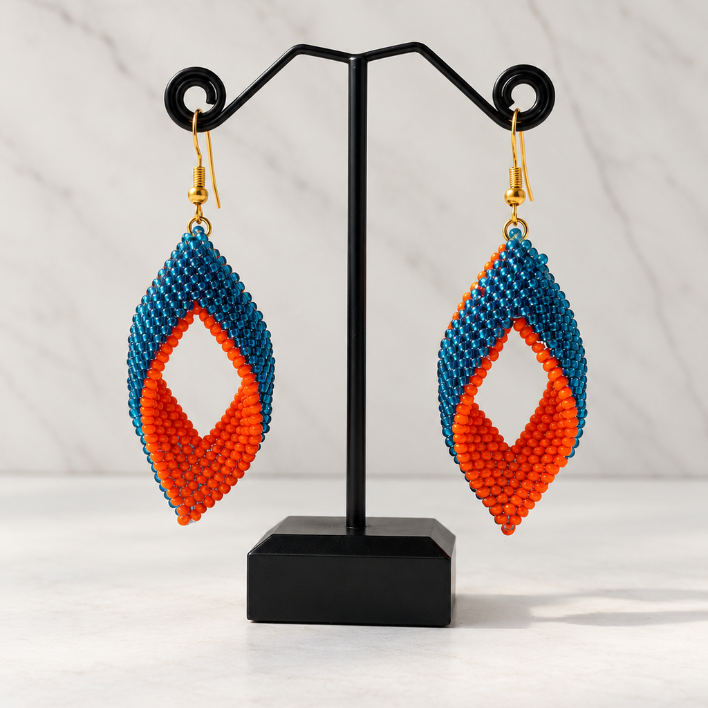 A pair of handmade beaded dangle earrings displayed on a black stand. The earrings feature a teardrop-shaped design with a blue top section, a white diamond-shaped center cutout, and an orange bottom section, finished with gold hooks.