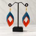 A pair of handmade beaded dangle earrings displayed on a black stand. The earrings feature a teardrop-shaped design with a blue top section, a white diamond-shaped center cutout, and an orange bottom section, finished with gold hooks.
