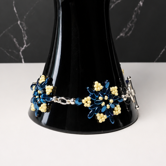 A beaded bracelet with blue and yellow floral motifs and silver chain details, displayed on a glossy black hand-shaped jewelry stand against a dark marble-patterned background.
