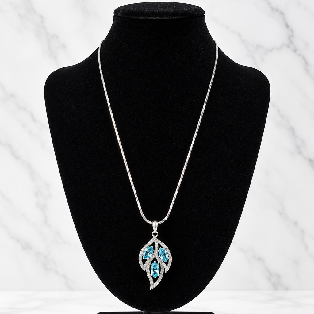 A silver pendant necklace with a leaf-shaped design set with bright blue stones, displayed on a black velvet jewelry bust against a white marble background.