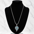 A silver pendant necklace with a leaf-shaped design set with bright blue stones, displayed on a black velvet jewelry bust against a white marble background.