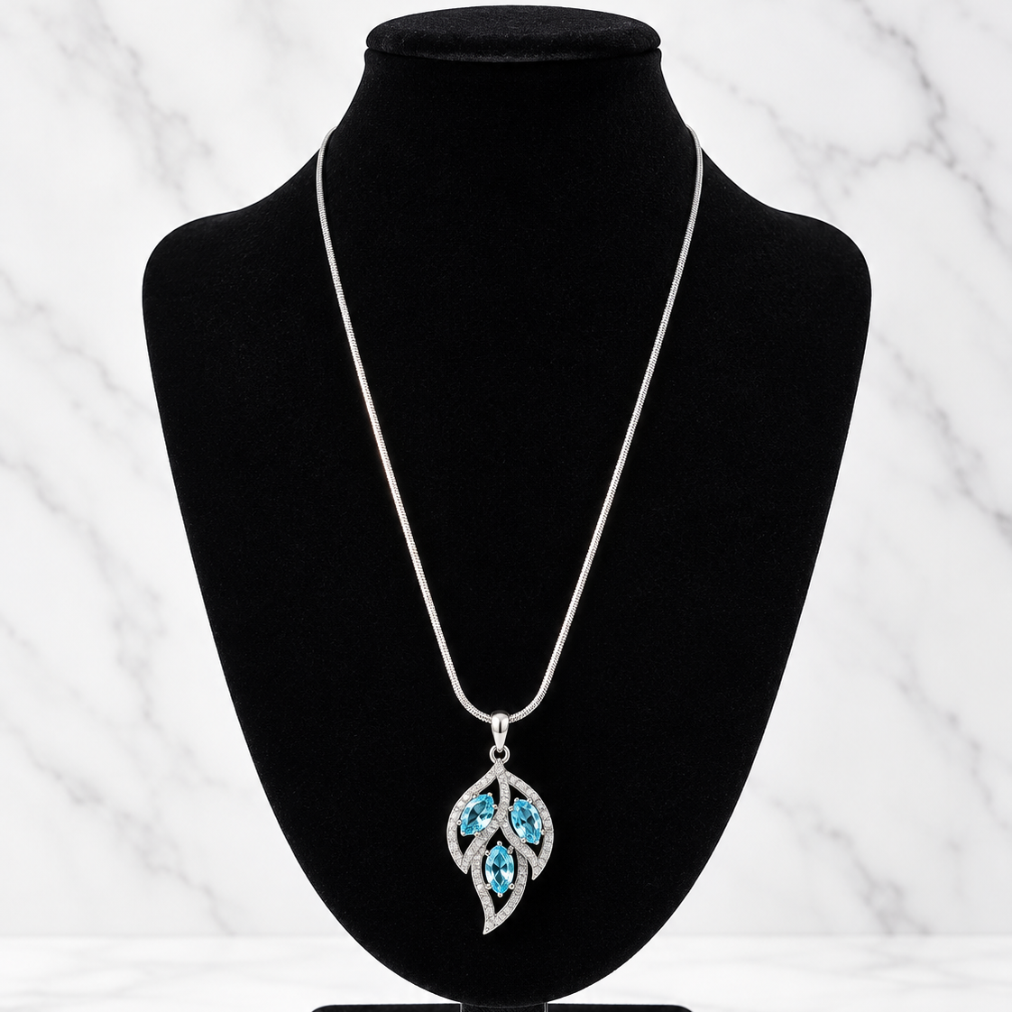 A silver pendant necklace with a leaf-shaped design set with bright blue stones, displayed on a black velvet jewelry bust against a white marble background.