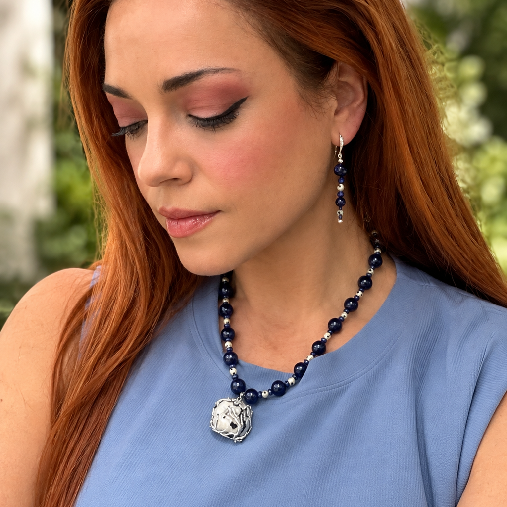 A woman with long red hair wearing a navy blue beaded necklace with a silver lion pendant and matching dangling earrings, paired with a light blue sleeveless top.