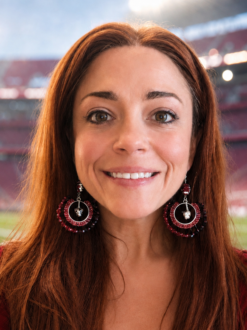 A smiling woman with long red hair wearing large circular dangling earrings and a red top, photographed in front of a blurred stadium background.
