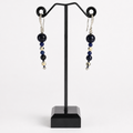 A pair of beaded drop earrings displayed on a black jewelry stand against a light gray background. The earrings feature dark navy blue and black round beads, small metallic silver beads, and silver hook ear wires.