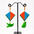 A pair of handmade beaded earrings displayed on a black jewelry stand against a white background. The earrings feature a diamond-shaped top with green beaded borders, split into blue and orange sections, and hang from gold-toned chains with green alligator charms below.