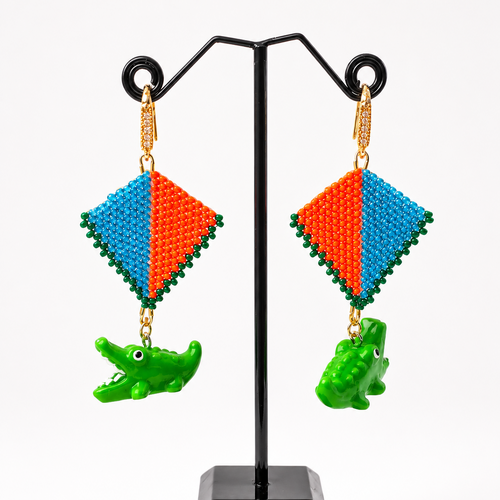 A pair of handmade beaded earrings displayed on a black jewelry stand against a white background. The earrings feature a diamond-shaped top with green beaded borders, split into blue and orange sections, and hang from gold-toned chains with green alligator charms below.