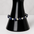 A beaded bracelet with dark blue, white, and black beads displayed on a glossy black hand-shaped jewelry stand against a light gray marble-patterned background.