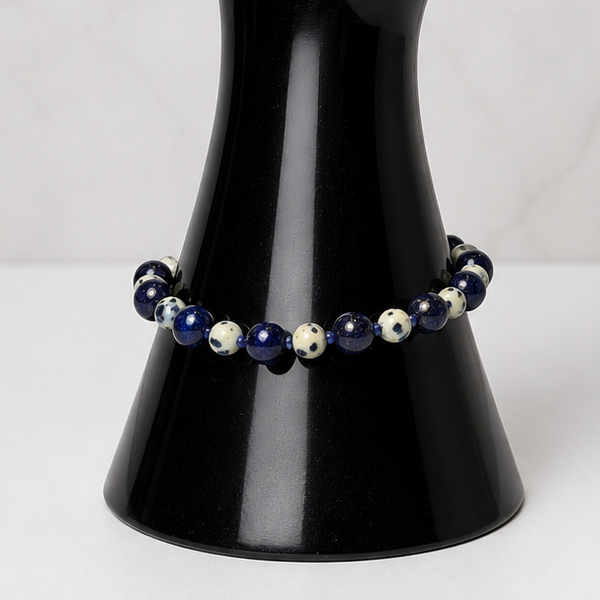 A beaded bracelet with dark blue, white, and black beads displayed on a glossy black hand-shaped jewelry stand against a light gray marble-patterned background.