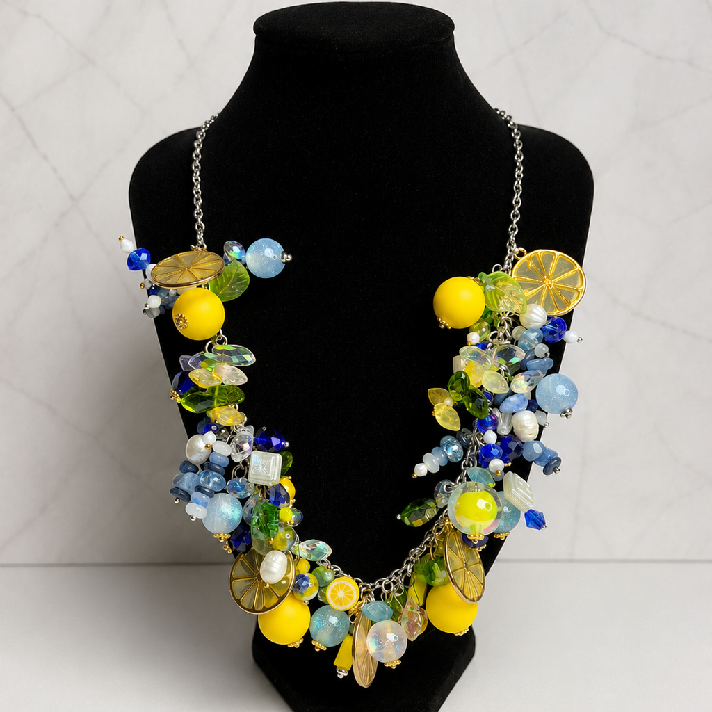 A colorful statement necklace with lemon and blue bead details, displayed on a black velvet jewelry bust against a light gray marble-patterned background.