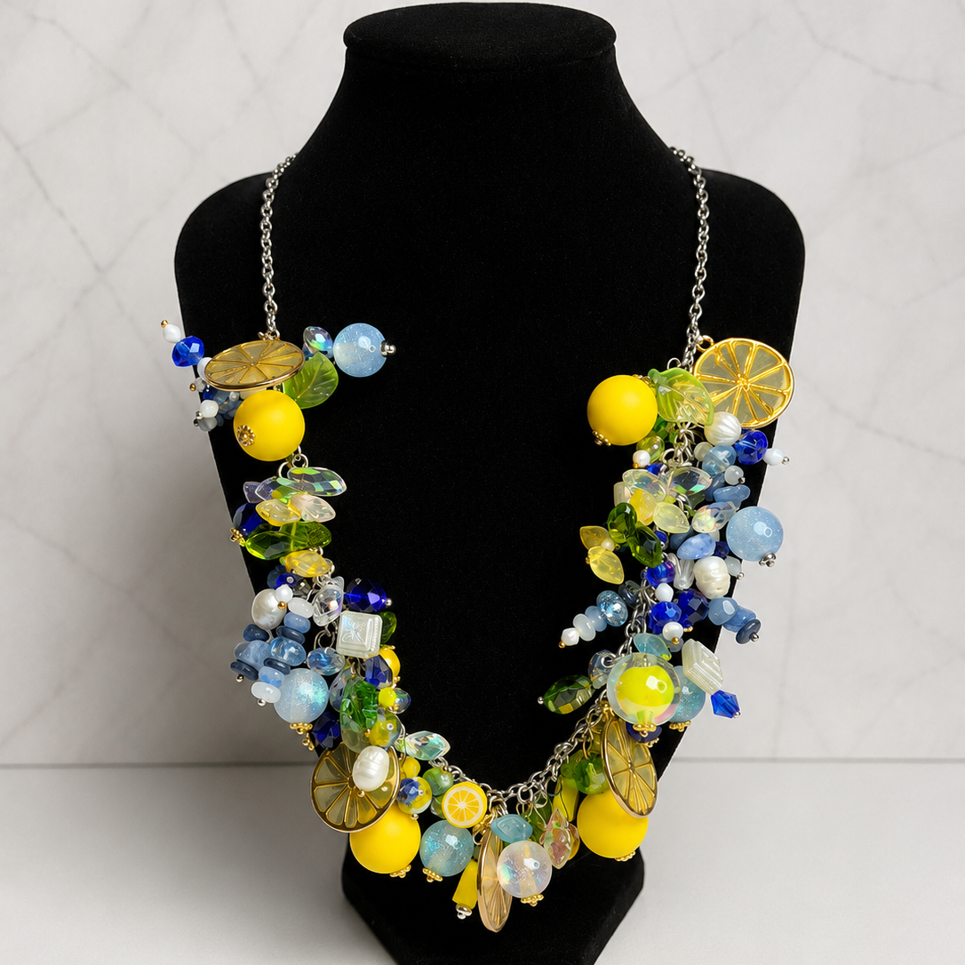 A colorful statement necklace with lemon and blue bead details, displayed on a black velvet jewelry bust against a light gray marble-patterned background.