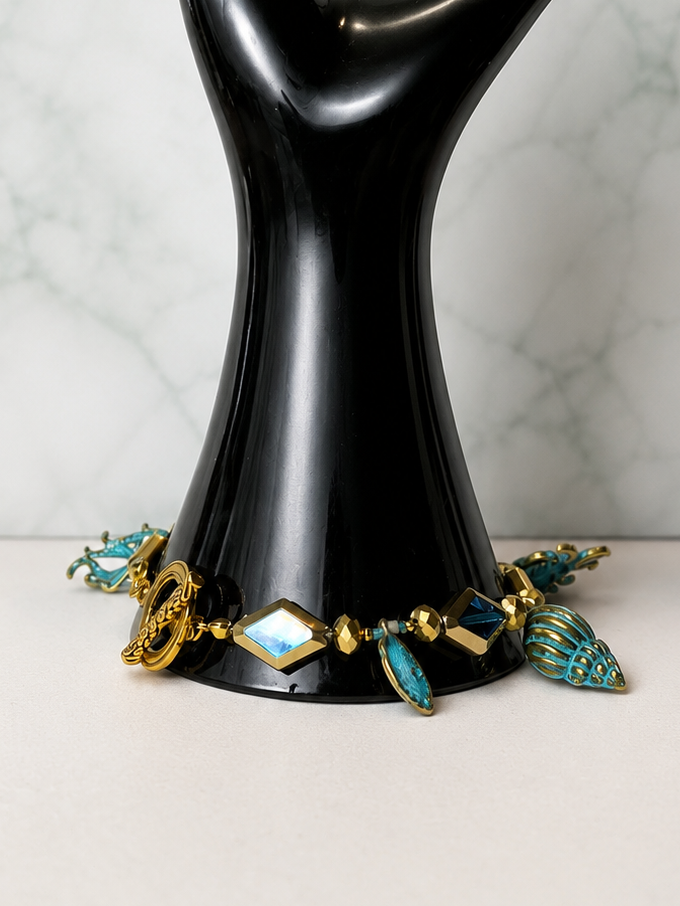 A gold-tone bracelet with turquoise and white stone accents displayed on a glossy black hand-shaped jewelry stand against a white marble-patterned background.