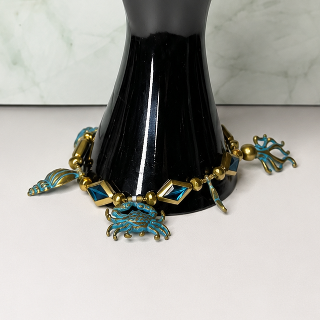 A black hand-shaped jewelry display stand holding a bracelet with turquoise blue and gold crab charms, set against a light marble-patterned background.