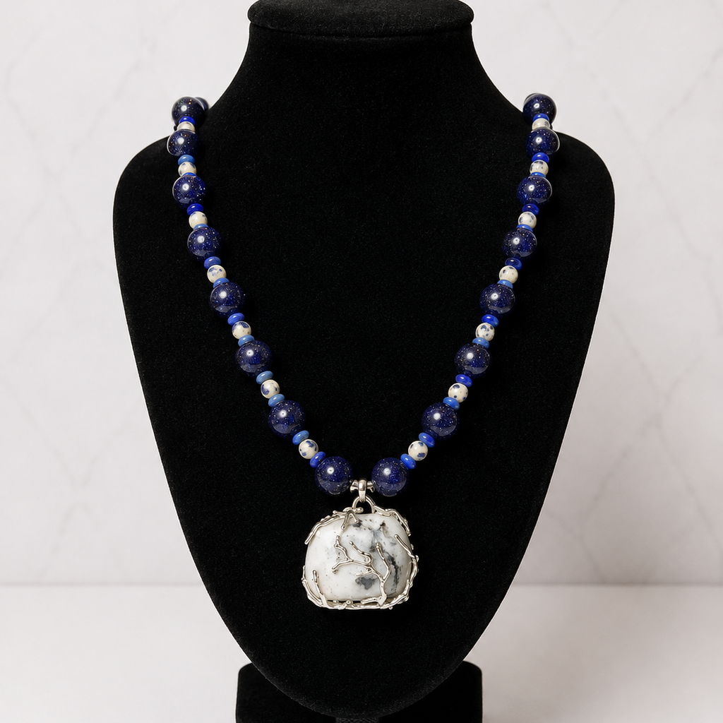A beaded necklace with deep blue and white beads, featuring a large white marble pendant with a silver border, displayed on a black velvet jewelry bust.