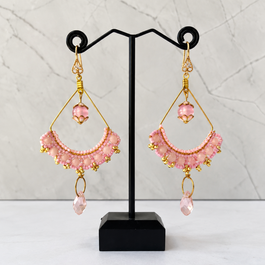A pair of handmade pink beaded dangle earrings with gold metal accents, displayed on a black jewelry stand against a light gray marble background.