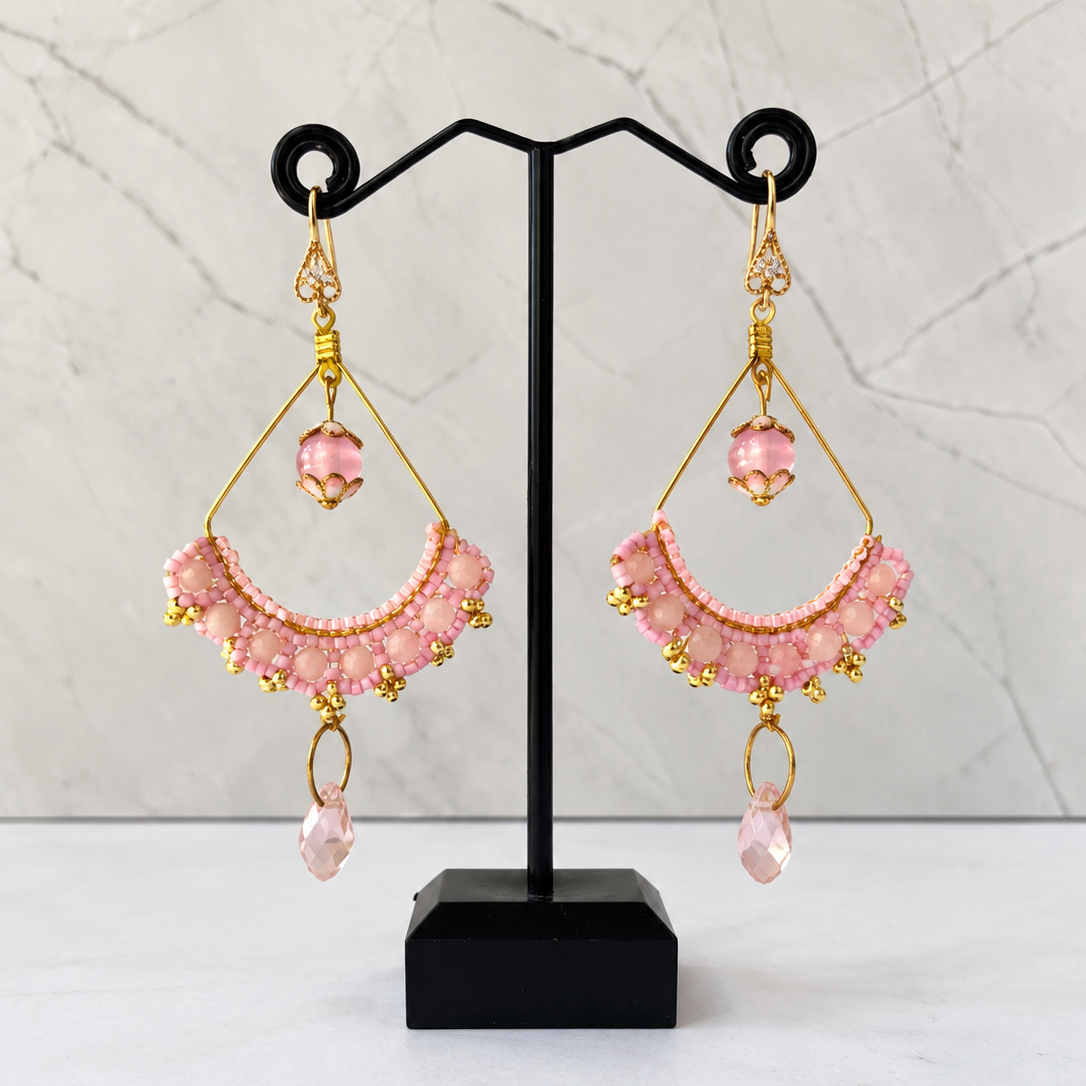 A pair of handmade pink beaded dangle earrings with gold metal accents, displayed on a black jewelry stand against a light gray marble background.